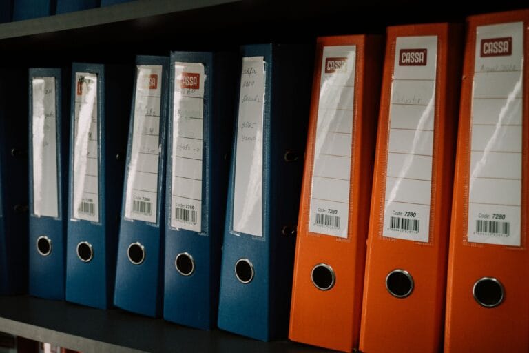Des classeurs et des dossiers papier rangés sur une étagère au-dessus d'un bureau.